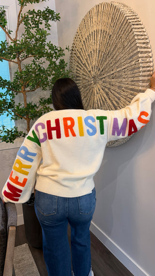 Person wearing a cream sweater with colorful 'Merry Christmas' text, standing in a room with decor elements.