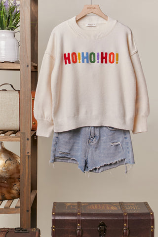 Beige sweater with 'HO HO HO!' text and denim shorts on a hanger against a neutral background.