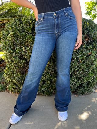 A woman wearing dark wash mid rise jeans with a navy stitched 7 on the back pocket. 