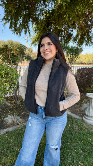 Woman wearing a black vest, beige sweater, and blue jeans standing outdoors.