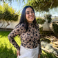 Woman standing outdoors in a garden wearing a patterned top and white pants.