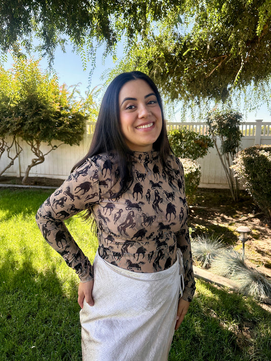Woman standing outdoors in a garden wearing a patterned top and white pants.