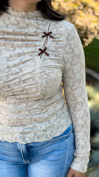 Close-up front view of beige long sleeve lace top showing ruched bodice, mock neck, and mini brown bow details