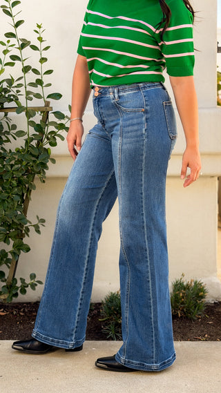 Woman wearing medium wash high rise wide leg Risen jeans with front seam detail and double copper buttons, styled with a green striped knit top and black western boots