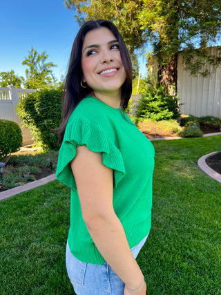 A woman wearing a green short sleeve ruffle sweater paired with blue jeans, standing outdoors.