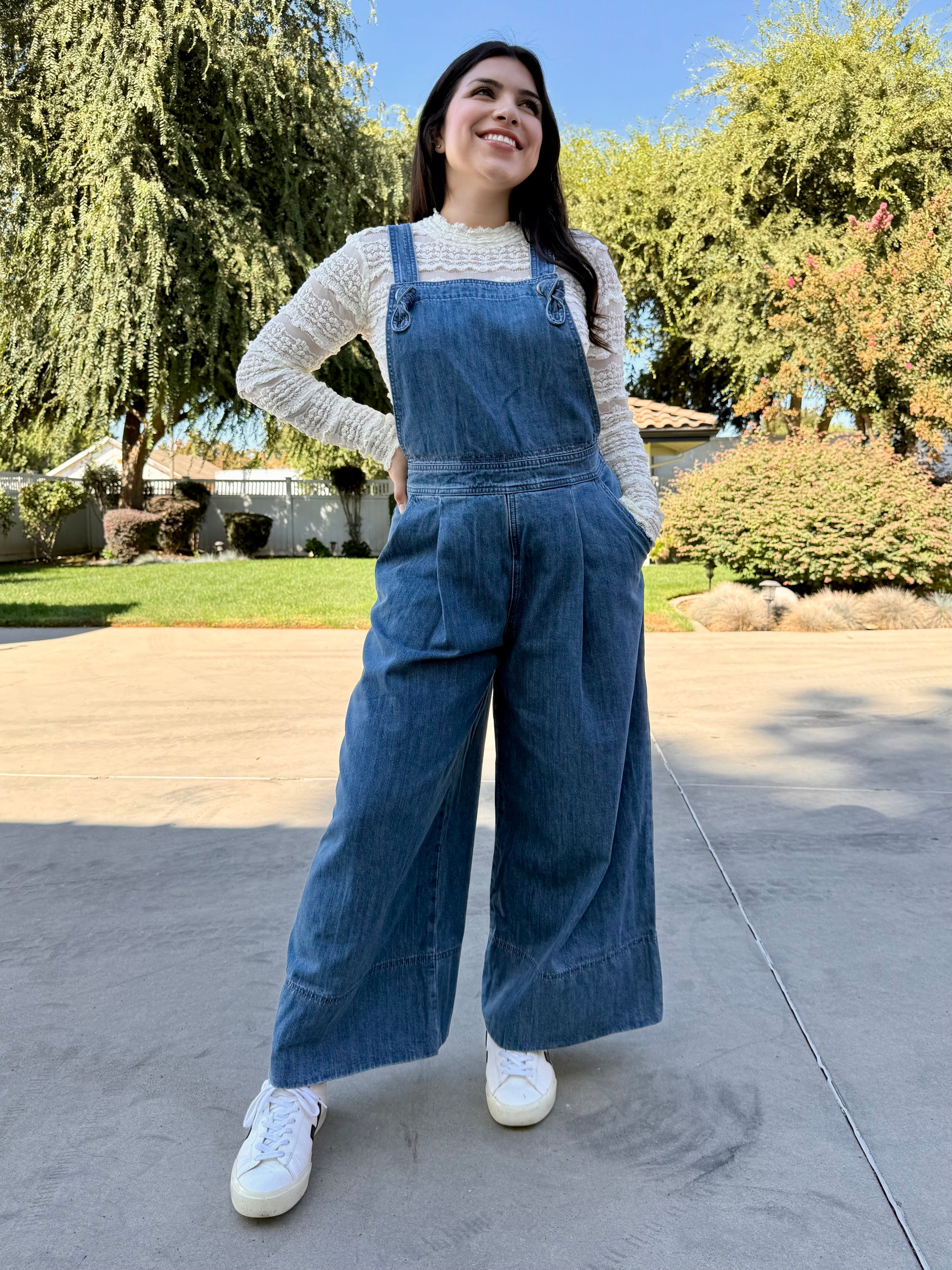 Woman wearing blue denim overalls with a white lace long sleeve and white sneakers standing 