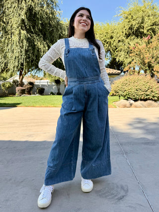 Woman wearing blue denim overalls with a white lace long sleeve and white sneakers standing 