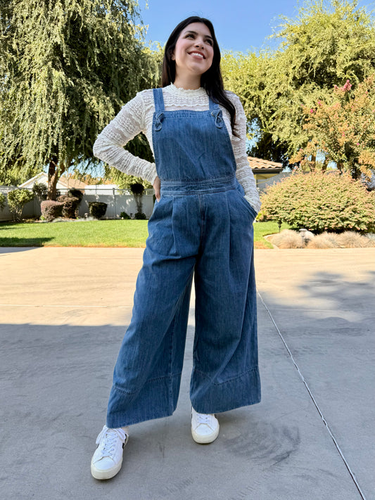 Woman wearing blue denim overalls with a white lace long sleeve and white sneakers standing 