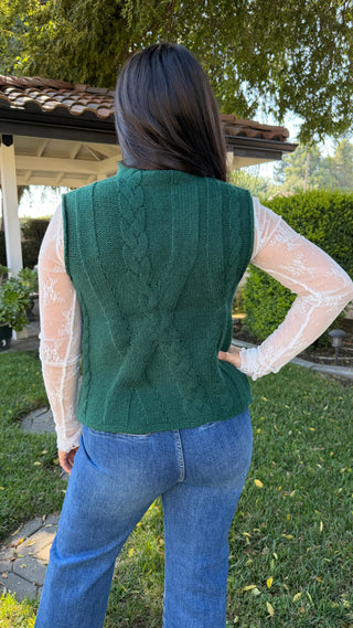 Woman wearing a green cable knit sleeveless sweater and blue jeans.