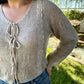 Woman wearing a beige cardigan and blue jeans standing outdoors with greenery in the background