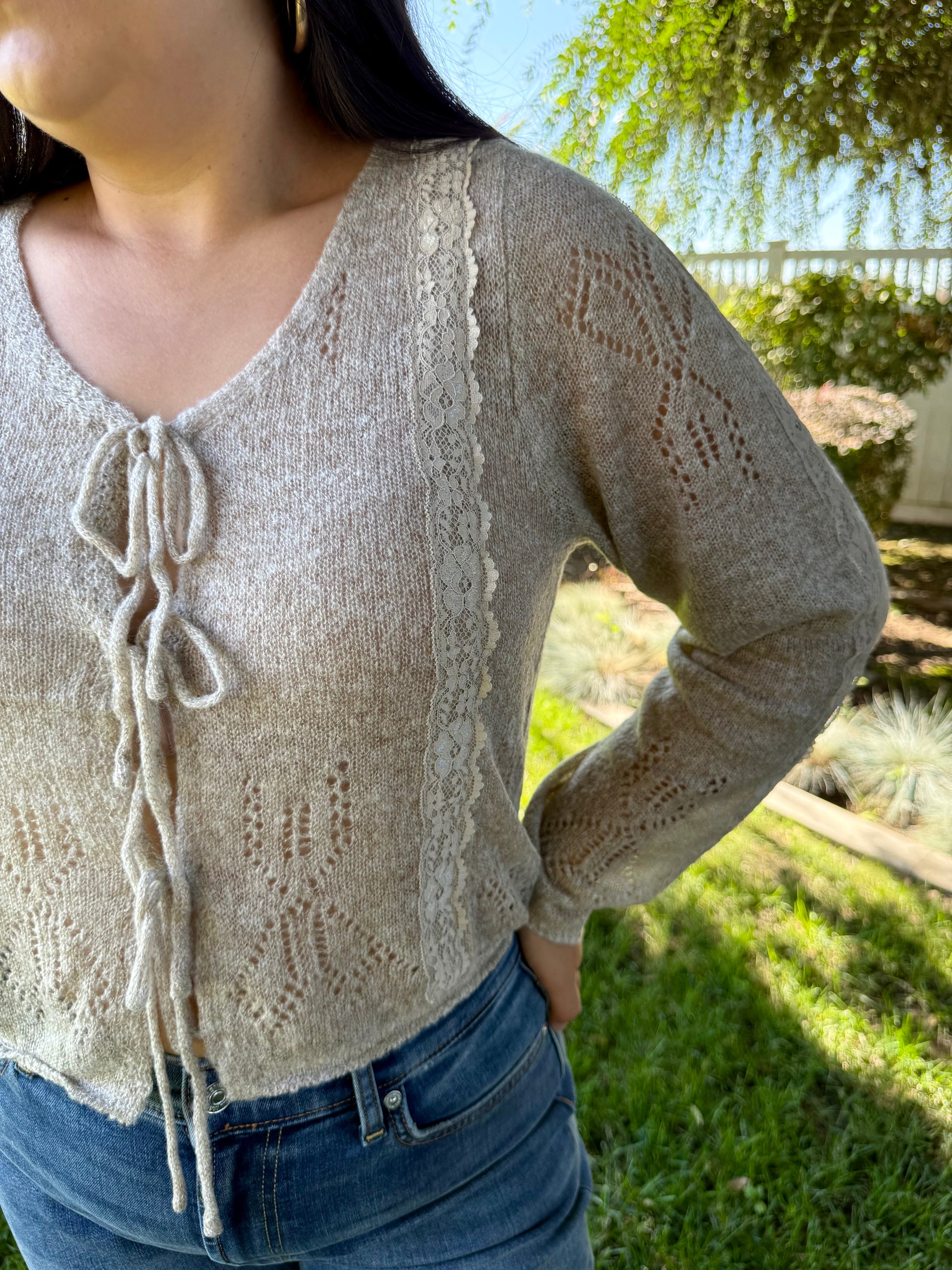 Woman wearing a beige cardigan and blue jeans standing outdoors with greenery in the background