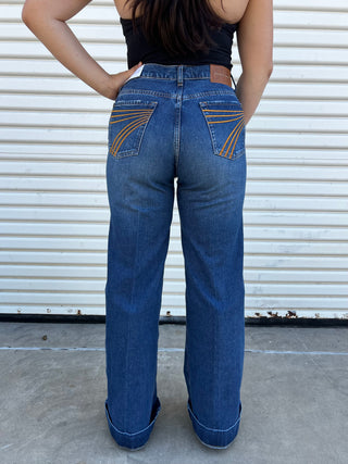 A woman wearing high waisted dark wash jeans with a gold 7 stitching on the back pocket. 