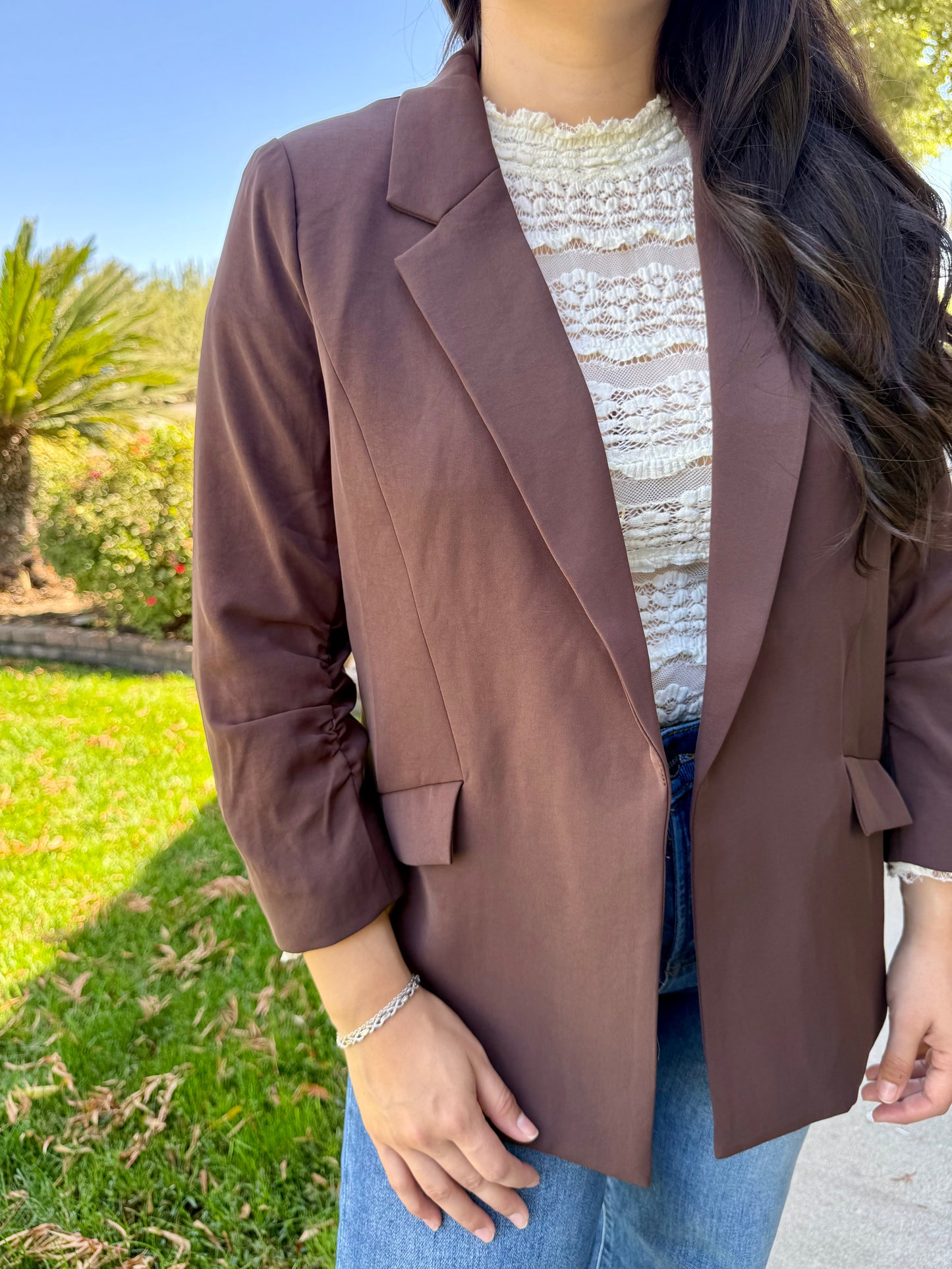 Person wearing a brown blazer over a white lace top with a blurred outdoor background