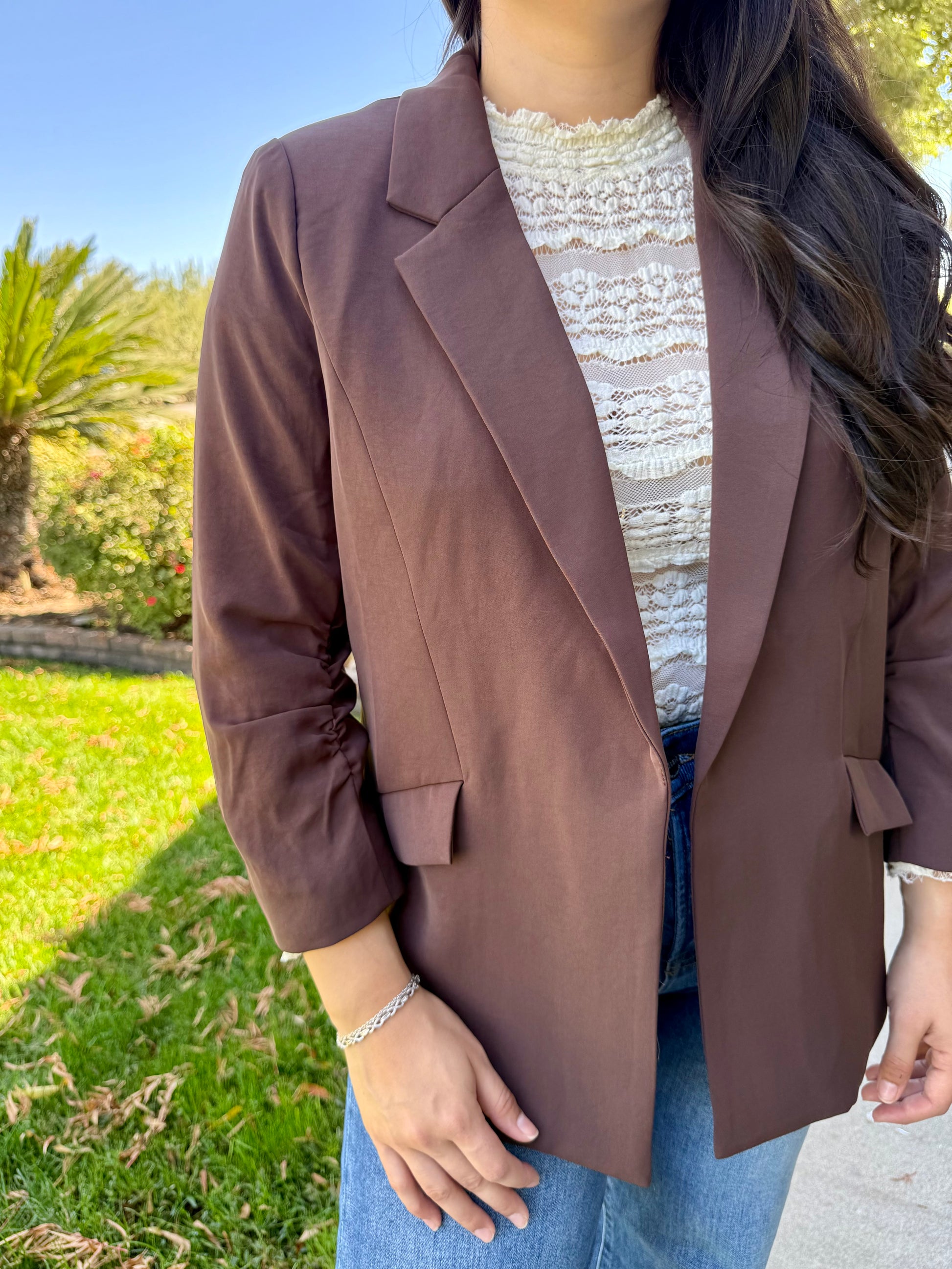 Person wearing a brown blazer over a white lace top with a blurred outdoor background