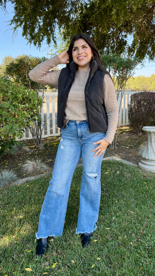 Woman standing outdoors wearing a beige sweater, black vest, and blue jeans.