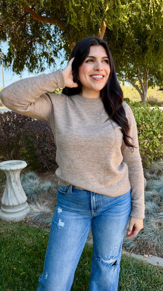 Woman wearing a beige sweater and blue jeans standing outdoors with greenery in the background