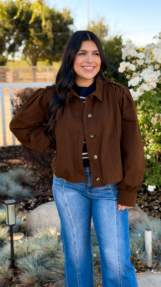 Woman wearing a brown cropped bomber jacket and blue jeans outdoors, front view