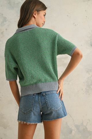 A woman wearing a short sleeve green and grey sweater with a collar while wearing blue jeans.