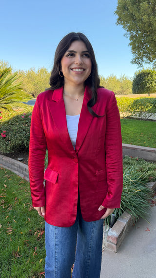Woman wearing a long sleeve red satin blazer and blue jeans.