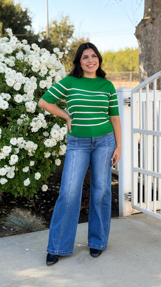 Woman wearing medium wash high rise wide leg Risen jeans with front seam detail and double copper buttons, styled with a green striped knit top and black western boots