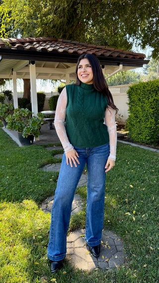 Woman wearing a green cable knit sleeveless sweater and blue jeans.