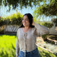 Woman wearing a beige cardigan and blue jeans standing outdoors with greenery in the background