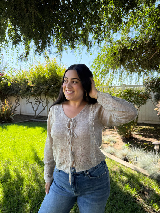 Woman wearing a beige cardigan and blue jeans standing outdoors with greenery in the background