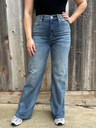 A woman wearing high waisted medium wash jeans in a wide leg style with a blue 7 stitching on the back pocket. 
