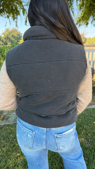 Person wearing a dark gray vest over a beige sweater with blue jeans outdoors.