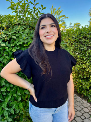 A woman wearing a black short sleeve ruffle sweater paired with blue jeans, standing outdoors.