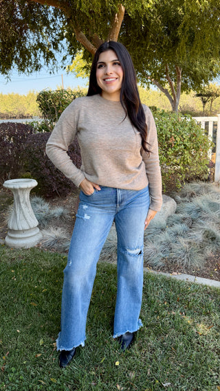 A woman wearing a long sleeve beige sweater with blue jeans outdoors.