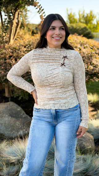 Smiling woman with hand on hip wearing a beige lace ruched mock neck long sleeve top with mini bows and light wash jeans outdoors