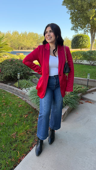 Woman wearing a long sleeve red satin blazer and blue jeans.