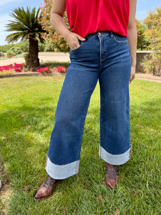A woman wearing high rise wide leg jeans with a big cuff on the bottom in a dark wash. 