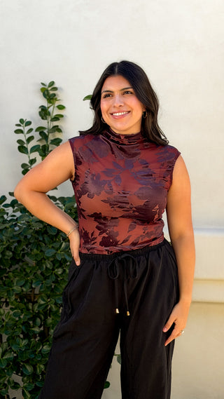 Front view of burgundy floral print sleeveless mock neck top, fitted and tucked into black wide leg drawstring pants