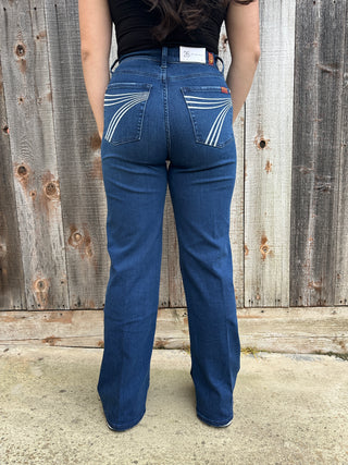 A woman wearing dark wash high waisted, wide leg jeans with a white 7 stitched on the back pocket. 