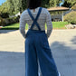 Woman wearing blue denim overalls with a white lace long sleeve and white sneakers standing 