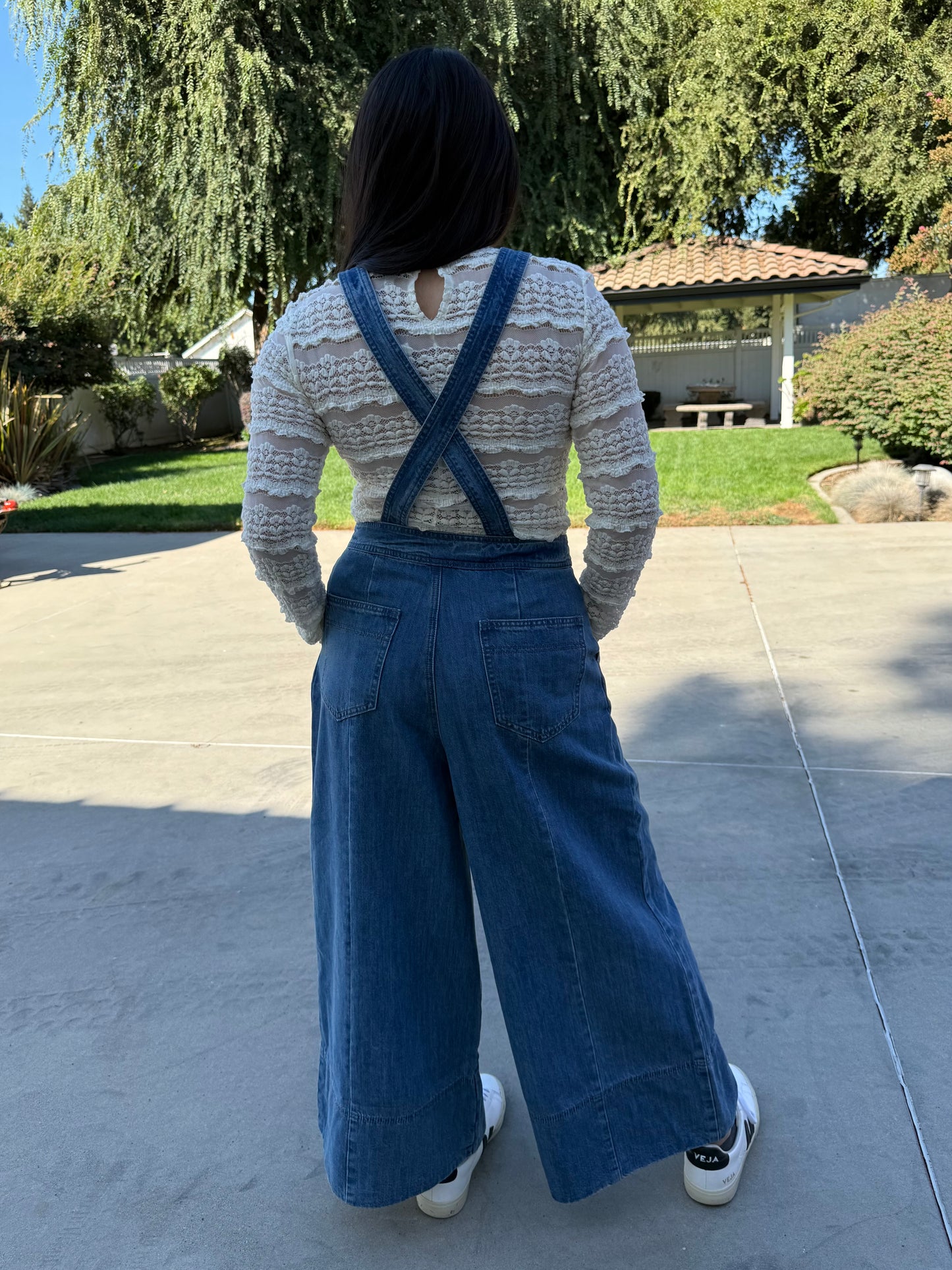 Woman wearing blue denim overalls with a white lace long sleeve and white sneakers standing 
