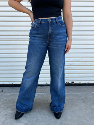 A woman wearing high waisted dark wash jeans with a gold 7 stitching on the back pocket. 