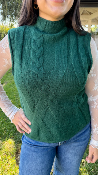 Woman wearing a green cable knit sleeveless sweater and blue jeans.