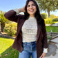 Woman posing outdoors in a residential area wearing a brown blazer, white lace top, and blue jeans.