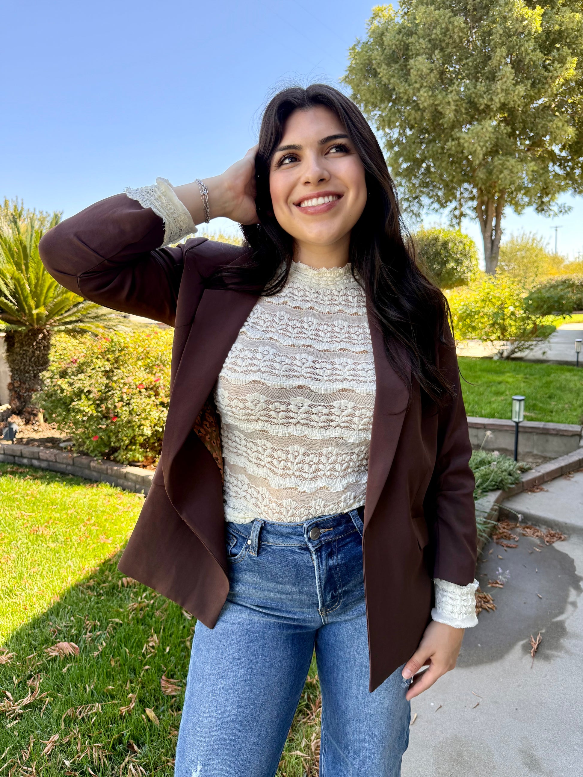 Woman posing outdoors in a residential area wearing a brown blazer, white lace top, and blue jeans.