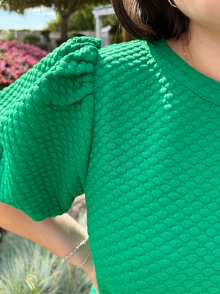 A woman wearing a green textured balloon sleeve top with a high neckline, paired with blue jeans.