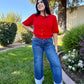 Woman in a red shirt and blue jeans standing on grass with trees and flowers in the background
