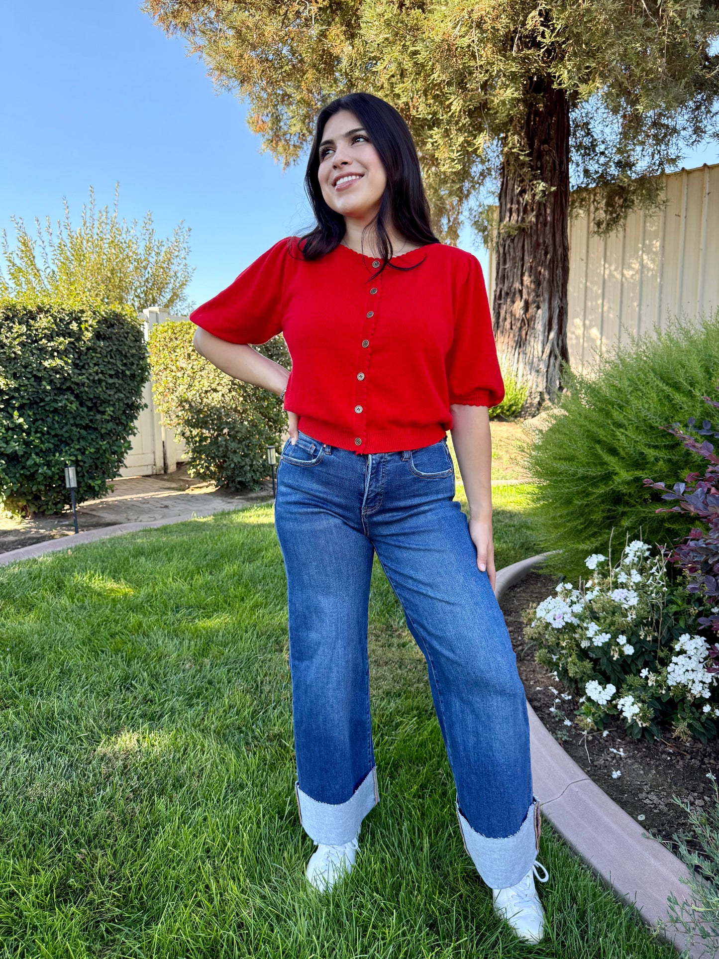 Woman in a red shirt and blue jeans standing on grass with trees and flowers in the background