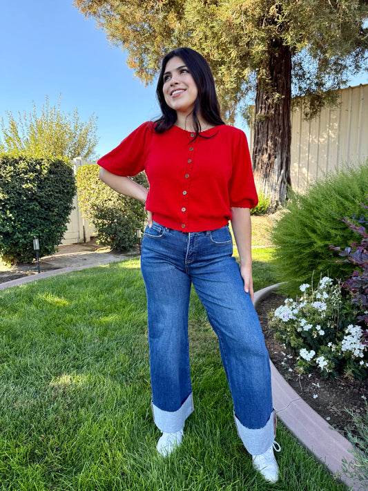 Woman in a red shirt and blue jeans standing on grass with trees and flowers in the background