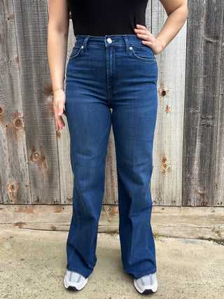 A woman wearing dark wash high waisted, wide leg jeans with a white 7 stitched on the back pocket. 