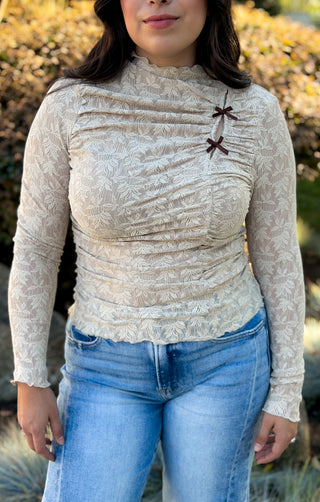 Woman wearing a beige lace ruched mock neck top with two mini brown bows at the shoulder, long sleeves with lettuce-edge cuffs, styled with light wash jeans