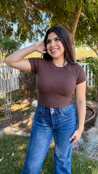 Woman wearing a brown top knitted top with a satin tie on the back and blue jeans.