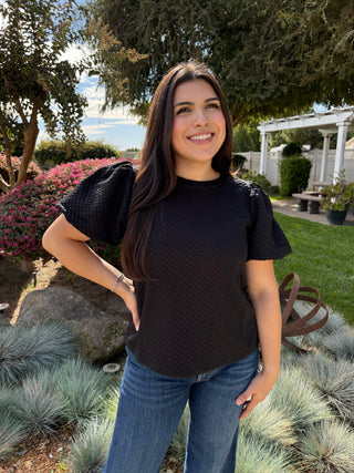 A woman wearing a black textured balloon sleeve top with a high neckline, paired with blue jeans.